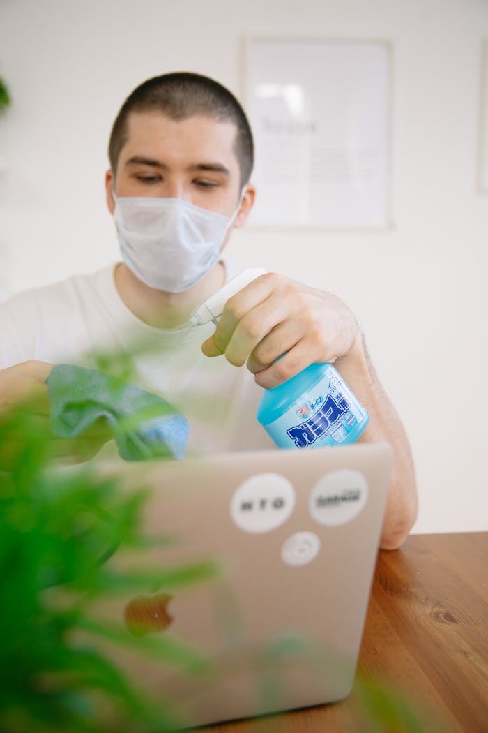 Adult man in face mask using spray bottle to sanitize laptop in an indoor setting.